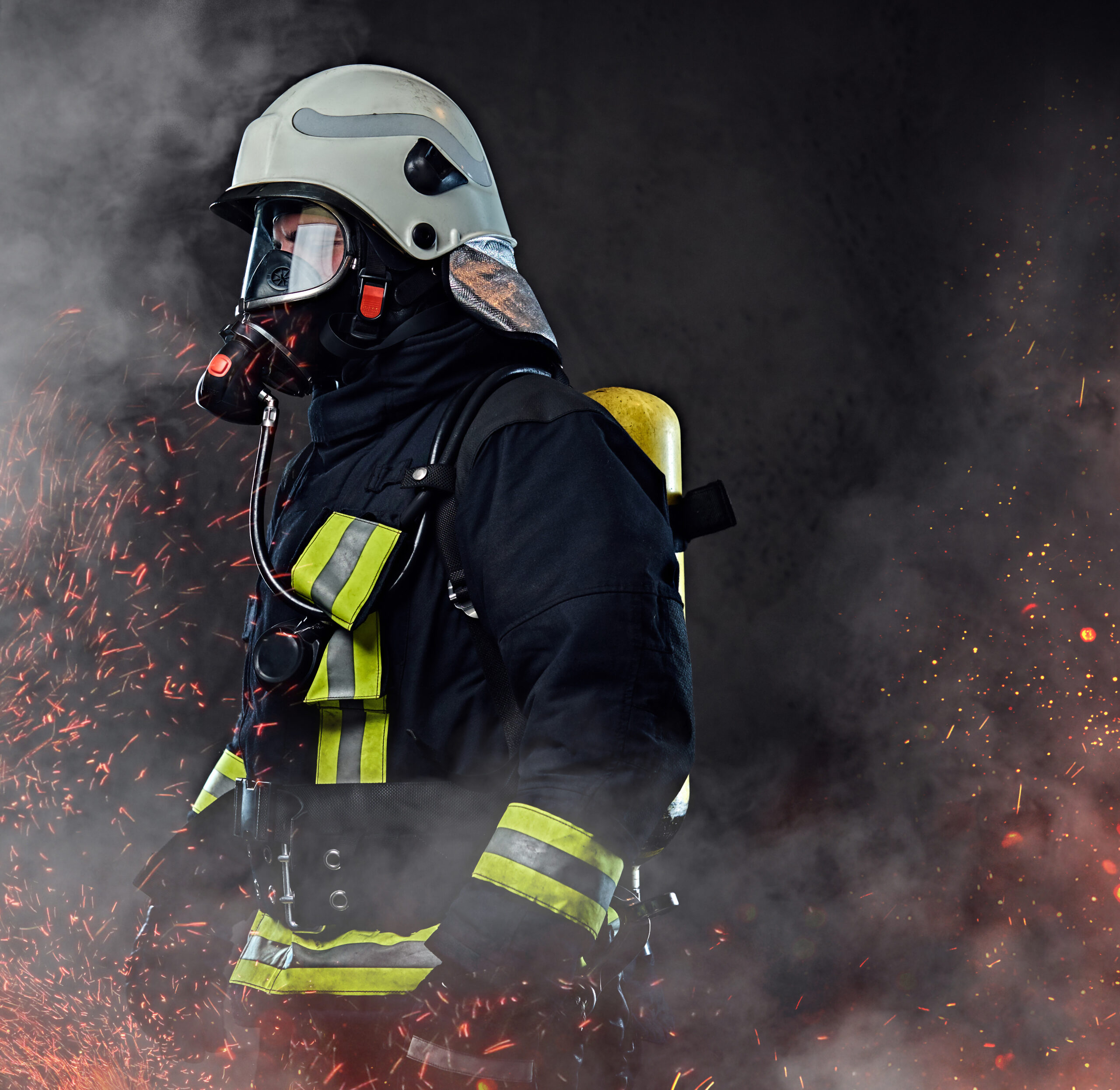 A professional firefighter dressed in uniform and an oxygen mask standing in fire sparks and smoke over a dark background.